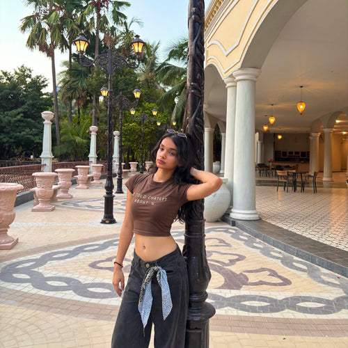 Woman in a brown baby tee with “i told chatgpt about u” text and dark pants standing in a decorative courtyard with palm trees and architectural columns.
