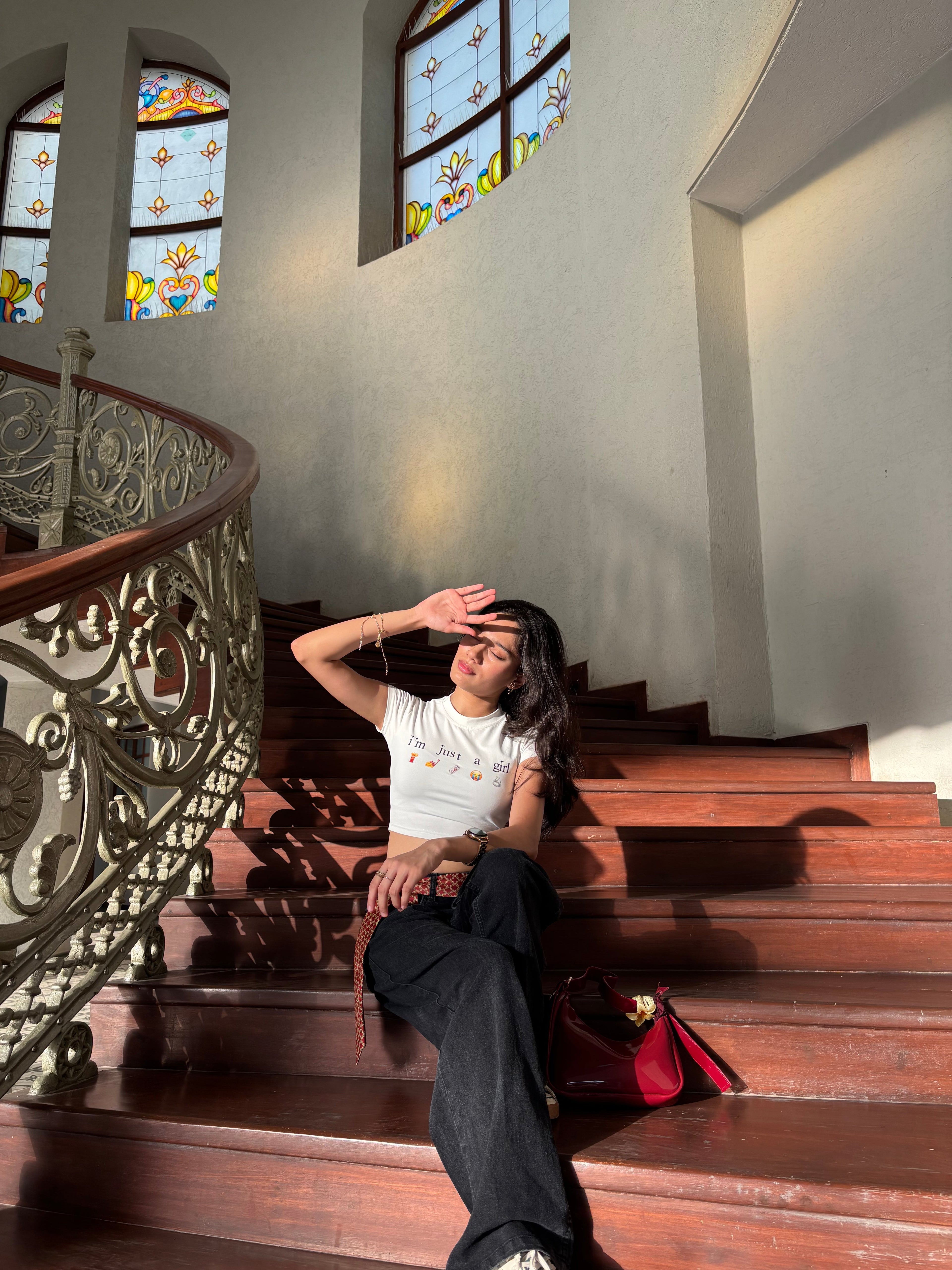 Woman sitting on a staircase in a building with stained glass windows wearing a cute baby fit crop top that says “i’m just a girl”