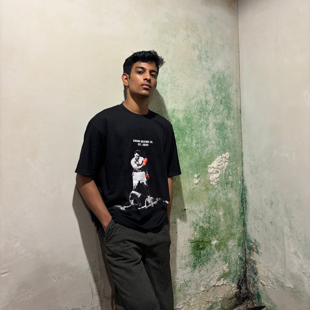 Person wearing a black t-shirt with a graphic design, standing against a wall with peeling paint.