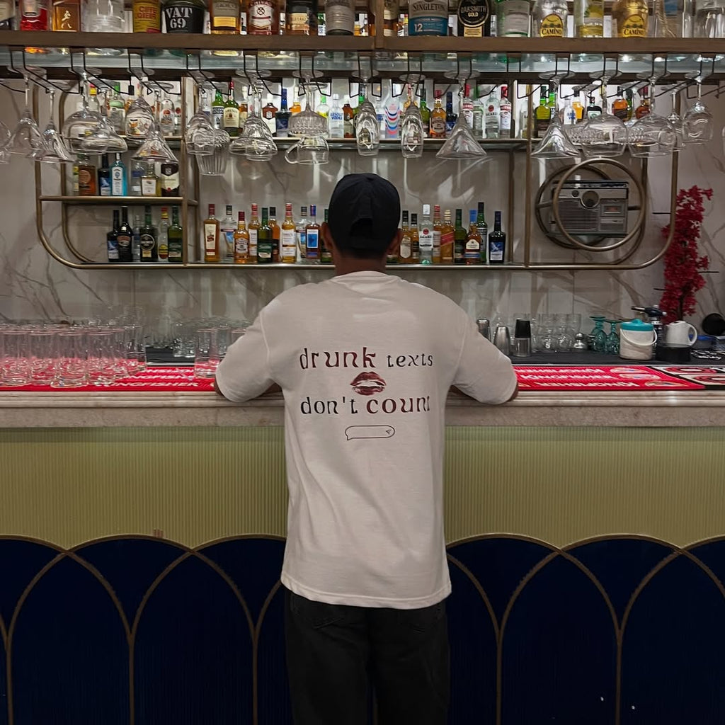 Person wearing a shirt with humorous text at a bar counter with drinks and glasses.