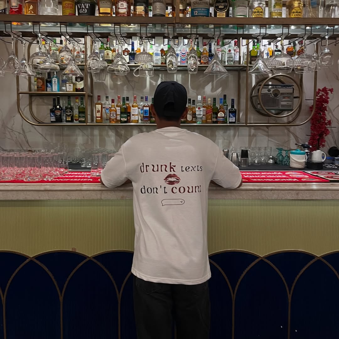 Person wearing a shirt with humorous text at a bar counter with drinks and glasses.