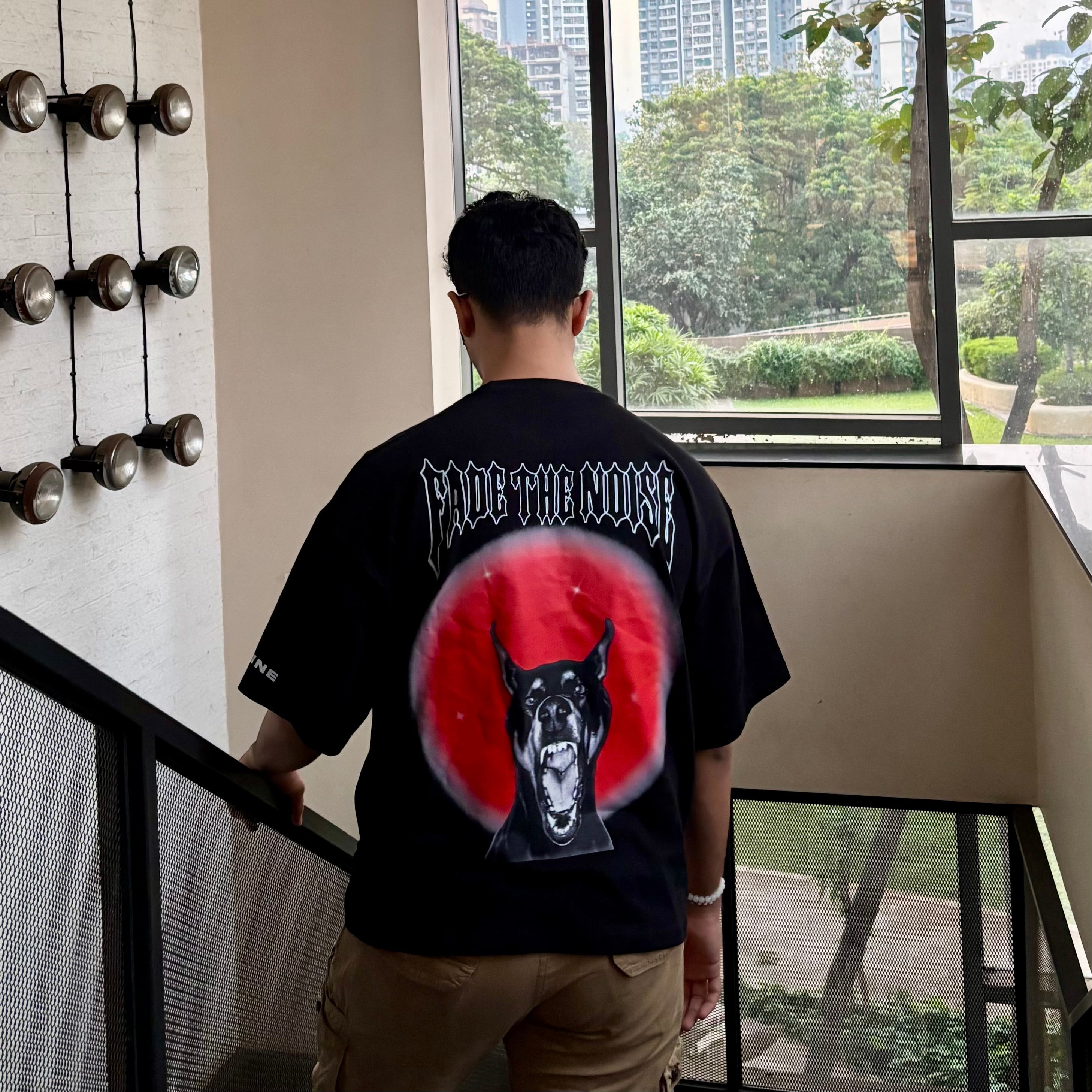 Person wearing a black t-shirt with graphic design and text, standing indoors near a window with a view of trees and buildings.