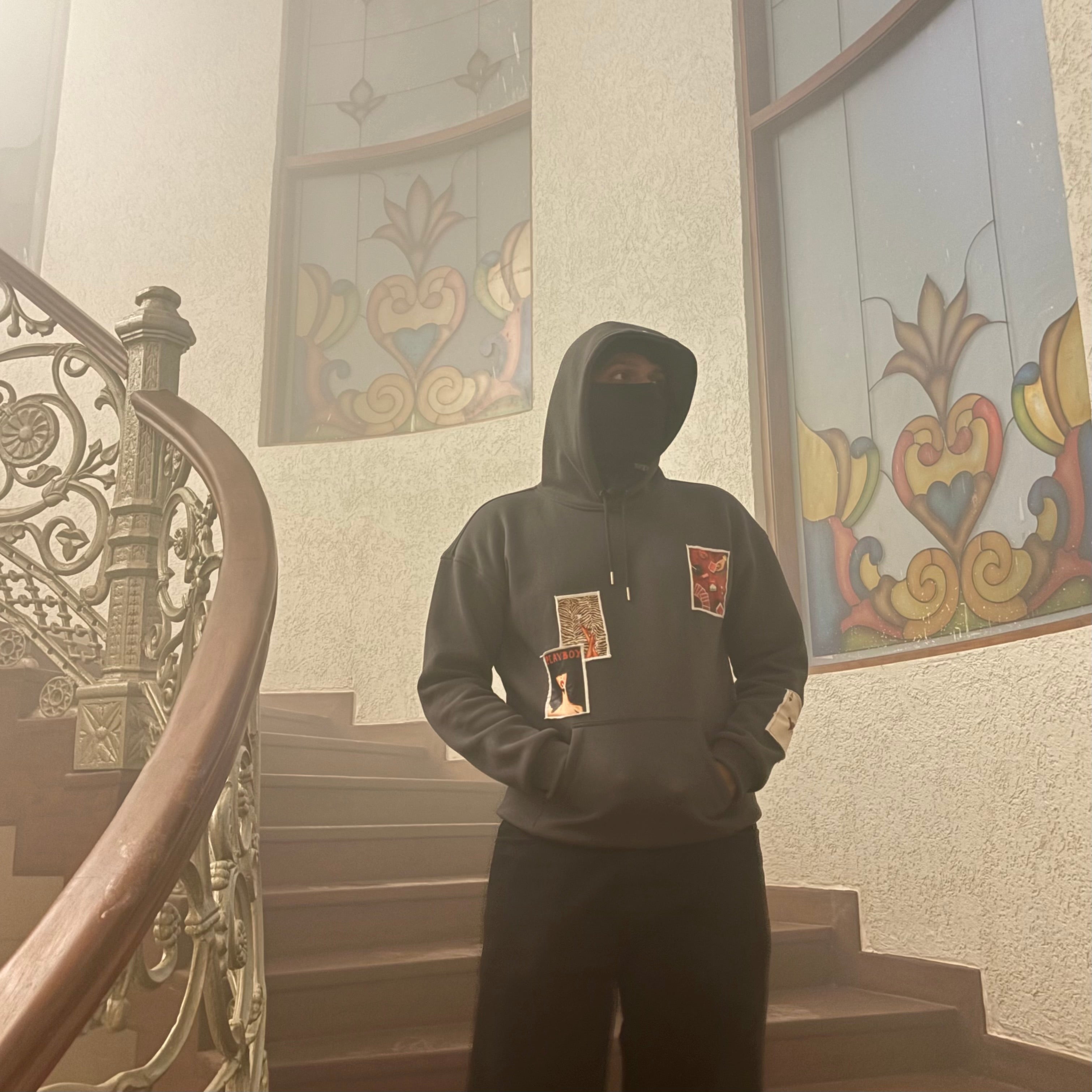 Person wearing a hoodie with patches in a foggy stairwell with decorative windows.