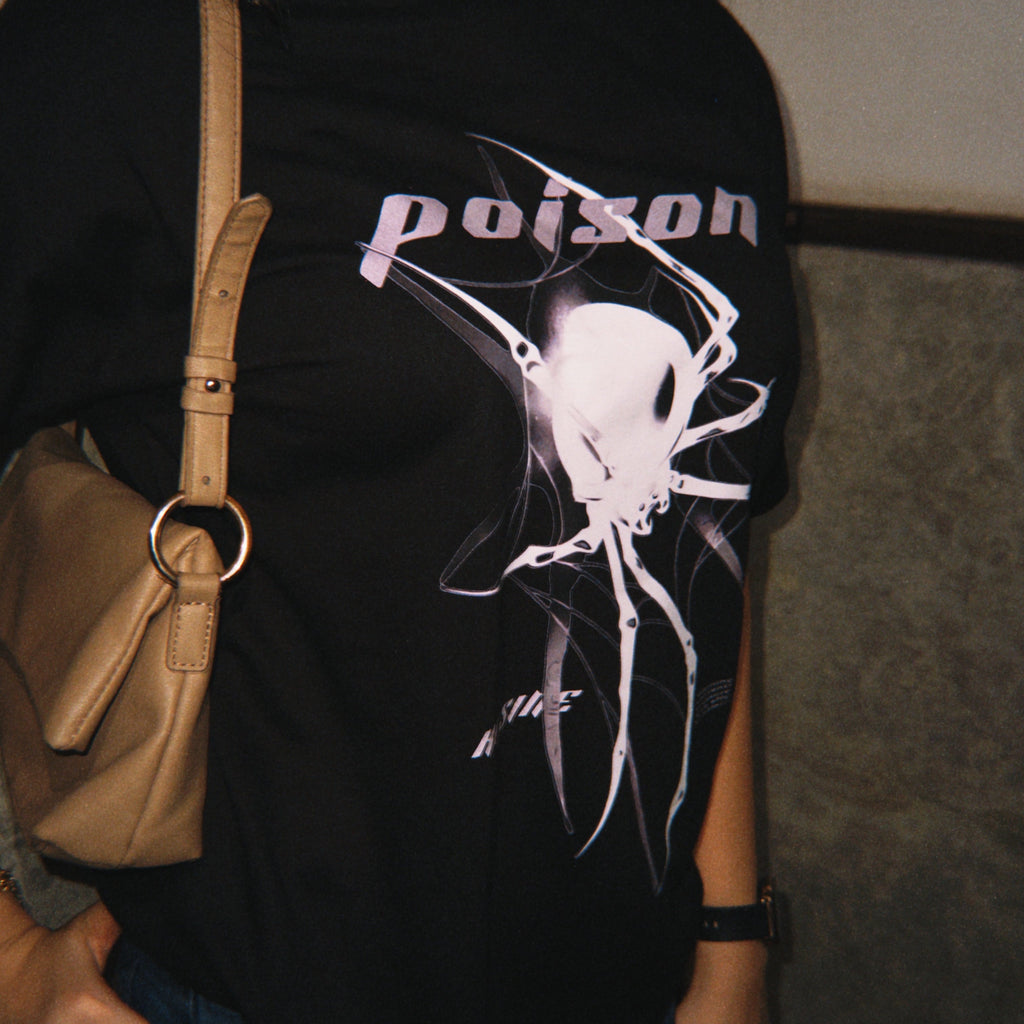 Person wearing a black t-shirt with a graphic design and the word 'poison', holding a beige handbag.