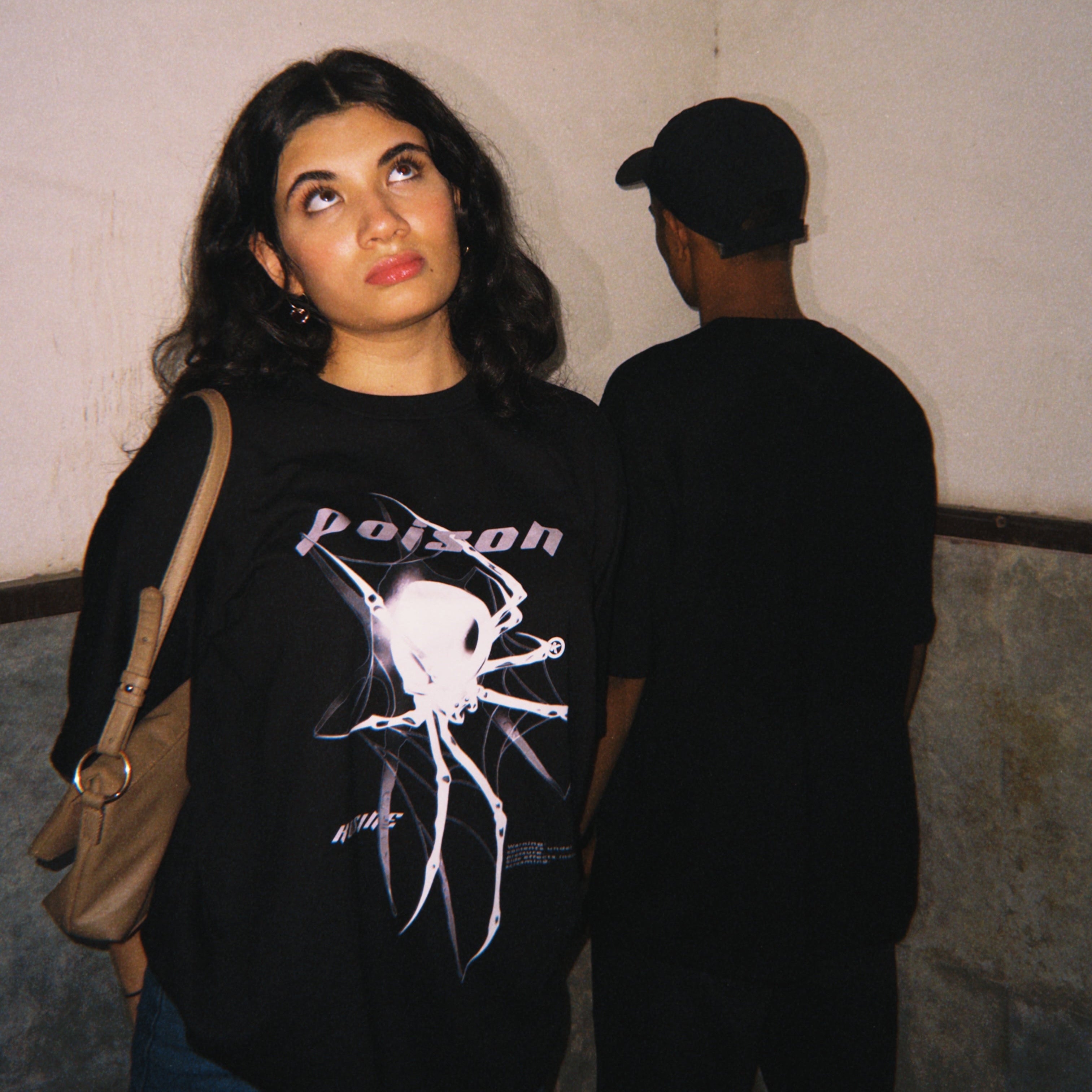 Woman wearing a black 'Poison' t-shirt with a man in the background.
