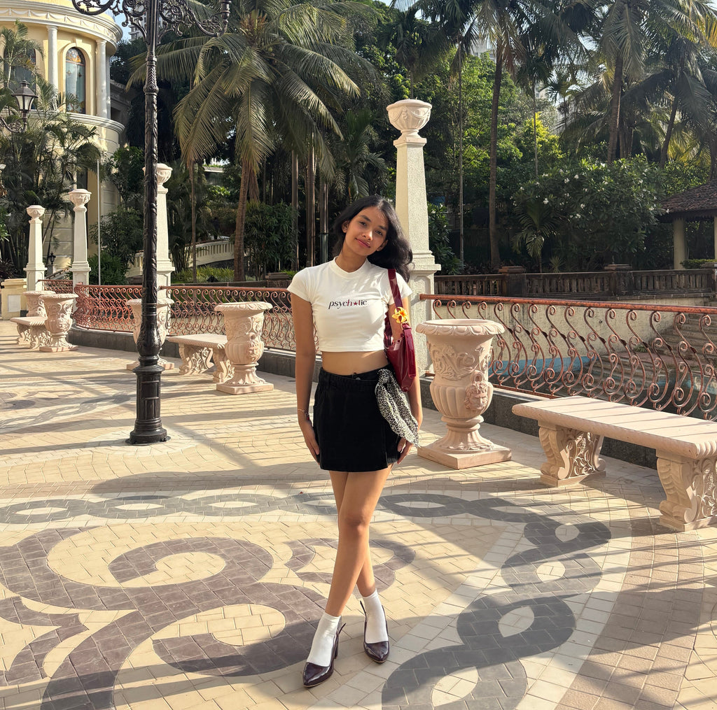 Woman wearing a cute baby-tee crop top that says “psychotic” and posing in a decorative outdoor setting with palm trees and architectural elements.