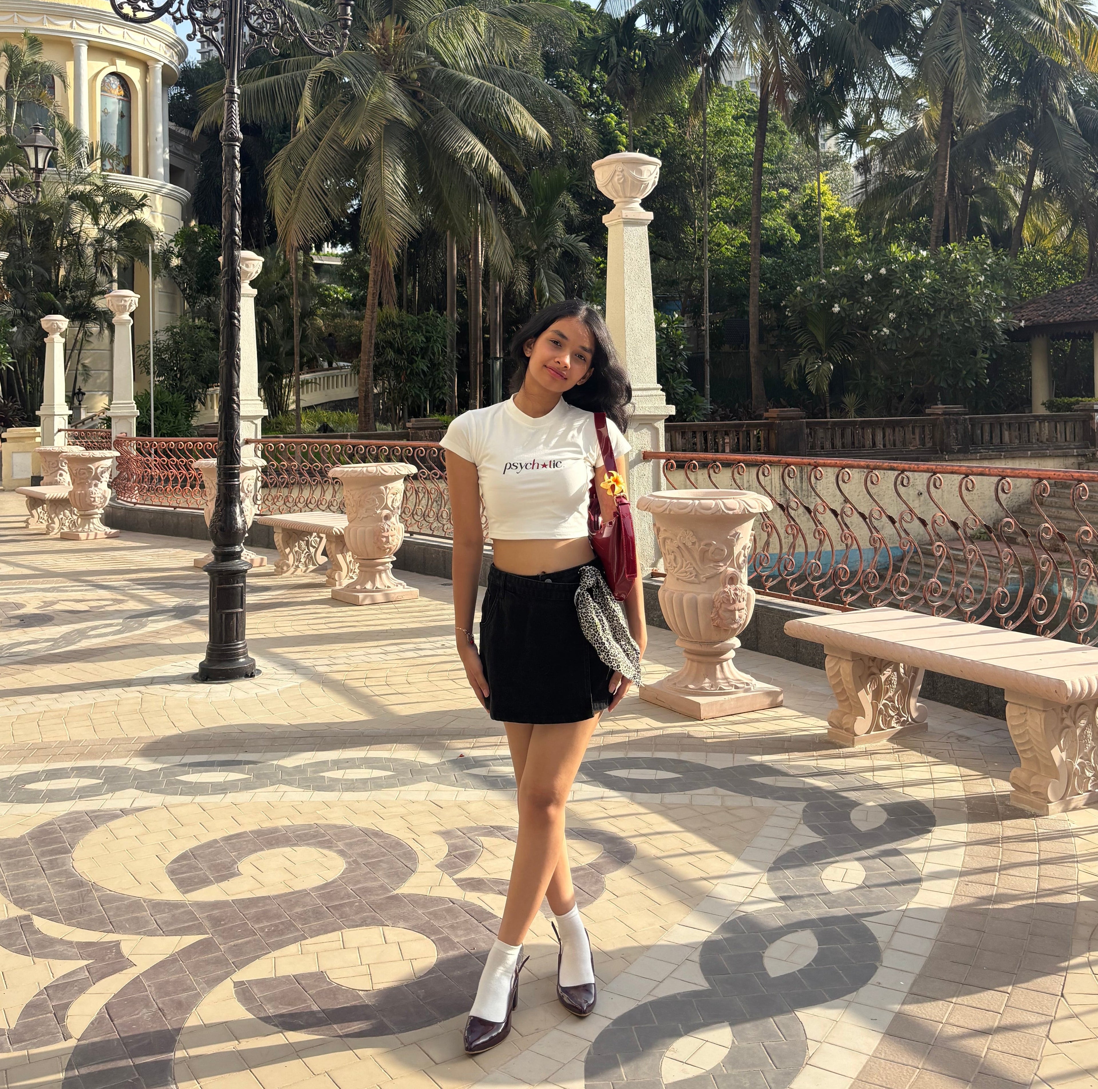 Woman wearing a cute baby-tee crop top that says “psychotic” and posing in a decorative outdoor setting with palm trees and architectural elements.