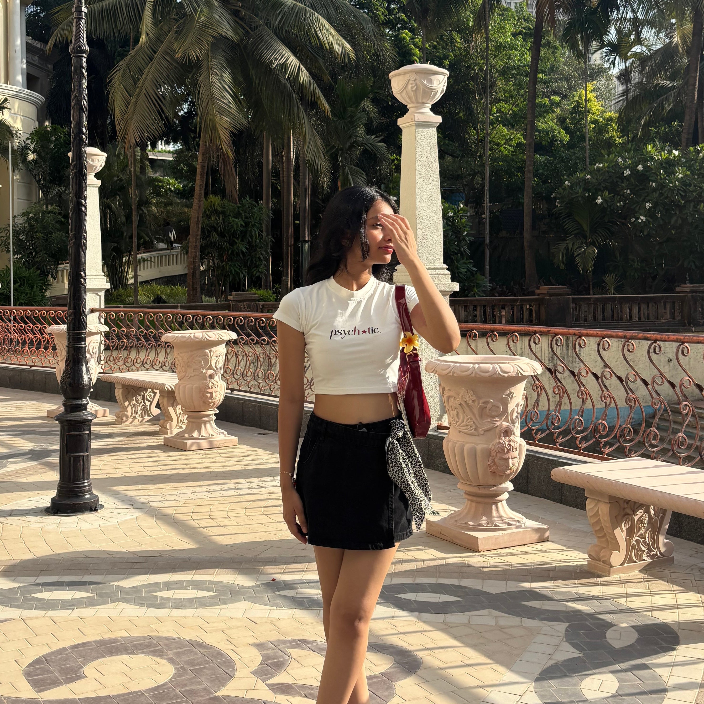 Woman wearing a cute crop top that says “psychotic” and standing in a park with palm trees and decorative columns.